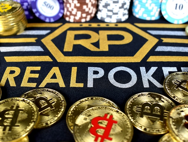 Stacks of poker chips in various colors are arranged in the background, while in the foreground, there are numerous golden coins featuring the Bitcoin symbol. The text 'REALPOKER' is prominently displayed in gold and silver lettering on a dark background.