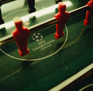 A foosball table is shown with red and black player figures positioned on the rods. The surface features the UEFA Champions League logo, indicating a themed table design.