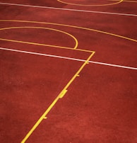 A sports court with a reddish-brown surface and bright yellow and white lines. The lines form geometric shapes, including rectangles and circles, suggesting markings for games like basketball or netball.