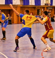 A dynamic indoor sports scene with players in motion on a court. Two athletes in blue and yellow uniforms are engaged in an intense handball game, with one player gripping the other's jersey. The background shows other players, several seated people, and advertisements on the walls.