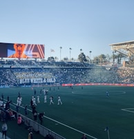 A large sports stadium filled with spectators. The field is occupied by soccer players, some walking and others scattered across the pitch. A massive screen displays a person, and flags fly on top of the stadium roof. The audience is dense, and the atmosphere seems lively with a visible banner showing 'BLUE WHITE & GOLD'.