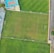 An aerial view of a sports complex featuring multiple green grass fields, including football pitches with marked goalposts. Adjacent to the fields is a blue-roofed building and a smaller area with a tennis or basketball court. Surrounding the complex, there's a road lined with trees and a dirt area with sparse vegetation.