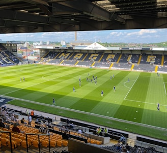 A sports stadium with a well-maintained grass field is depicted. Players are spread across the field, engaging in warm-up activities. The stands are partially filled with spectators, and the stadium is surrounded by urban buildings under a partly cloudy sky.