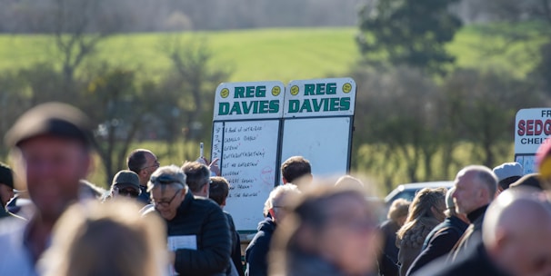 A group of people gathered outdoors with two large betting boards in the background labeled 'REG DAVIES'. The setting appears to be a countryside location with trees and fields visible in the distance. The crowd consists of individuals dressed in jackets and hats, engaging in conversation.