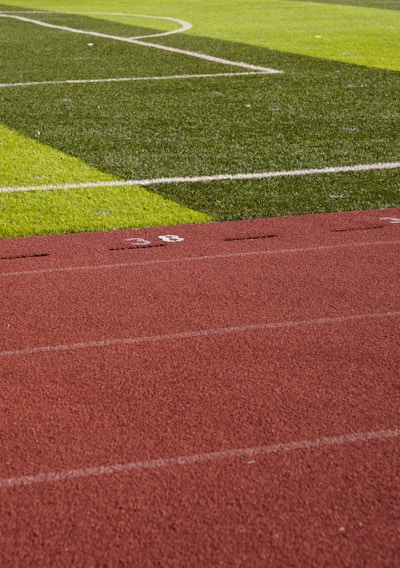 A sports field with vivid green grass divided into sections by white lines in the background. In the foreground, a track with a reddish-brown surface is marked with numbers and lines.