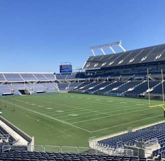 A large, empty sports stadium with blue seats surrounding a well-maintained green grass field. Bright sunlight creates clear shadows, and there are two goal posts on opposite ends of the field. A big electronic scoreboard displays some graphics, and the sky is clear and blue.